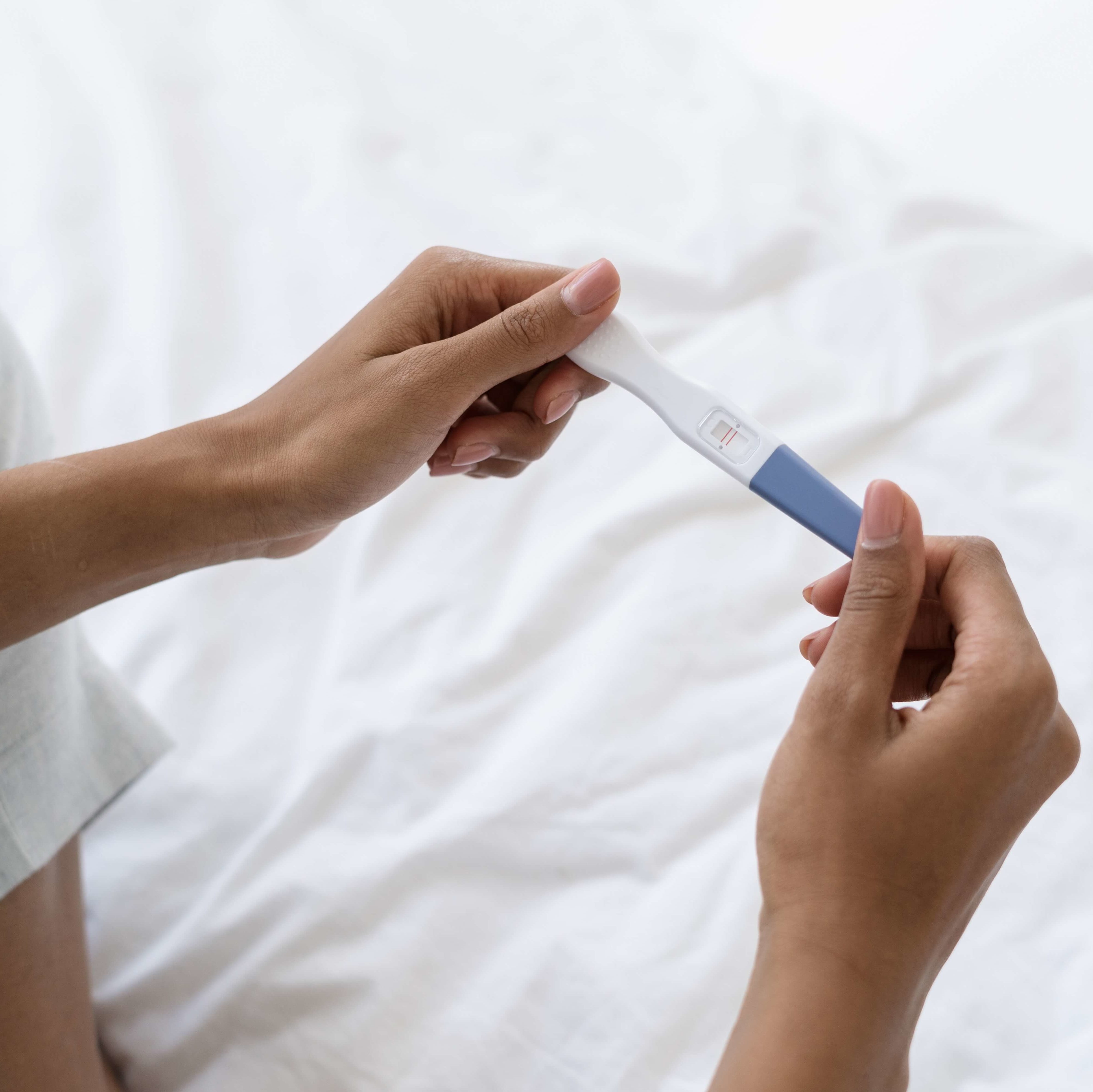 photo of a woman's hands holding a pregnancy test over a bed with white sheets